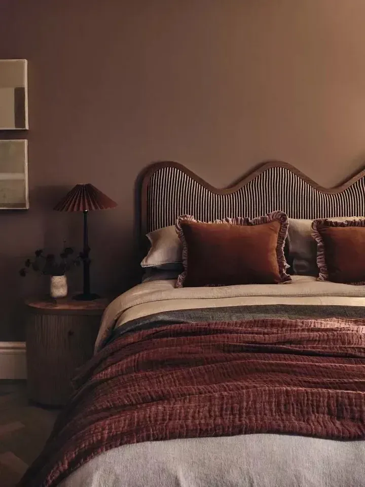 dark boho bedroom ideas with deep red bedding, curved headboard, dark lamp, layered pillows, and warm wall color