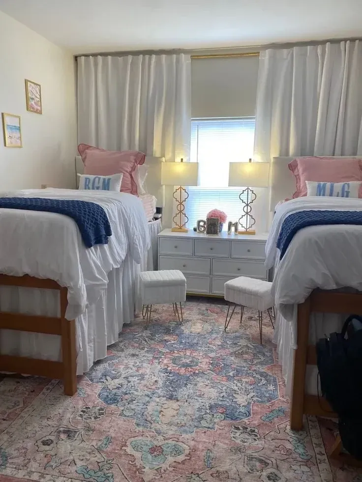 pink and blue dorm room ideas with blue throws, pink pillows, matching dorm beds, patterned rug, lamps, and white curtains