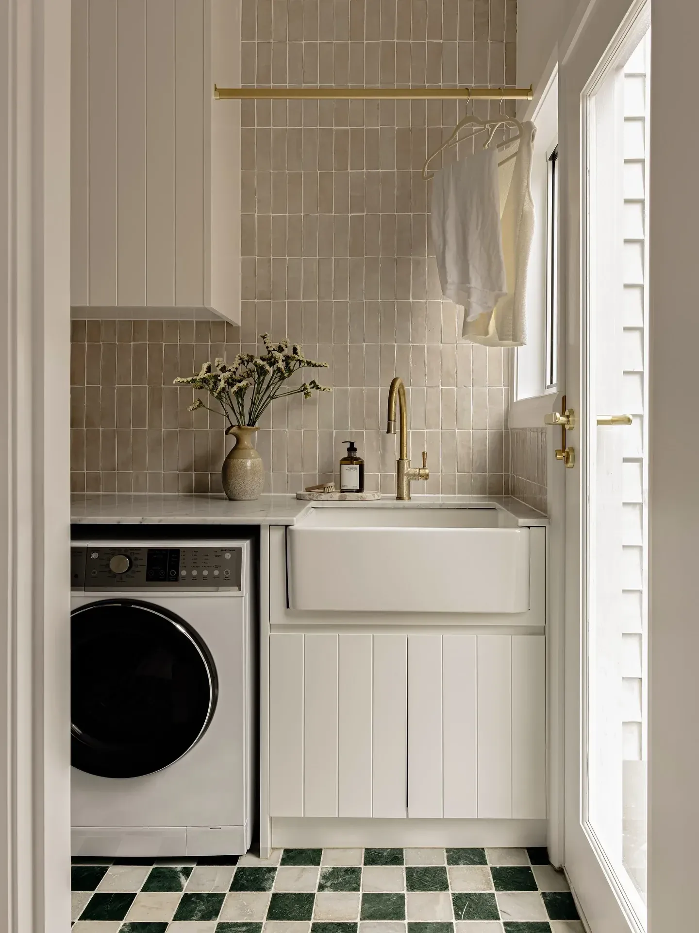 small laundry room ideas with brass hanging rail, farmhouse sink, washer, beige tile, and coat hangers