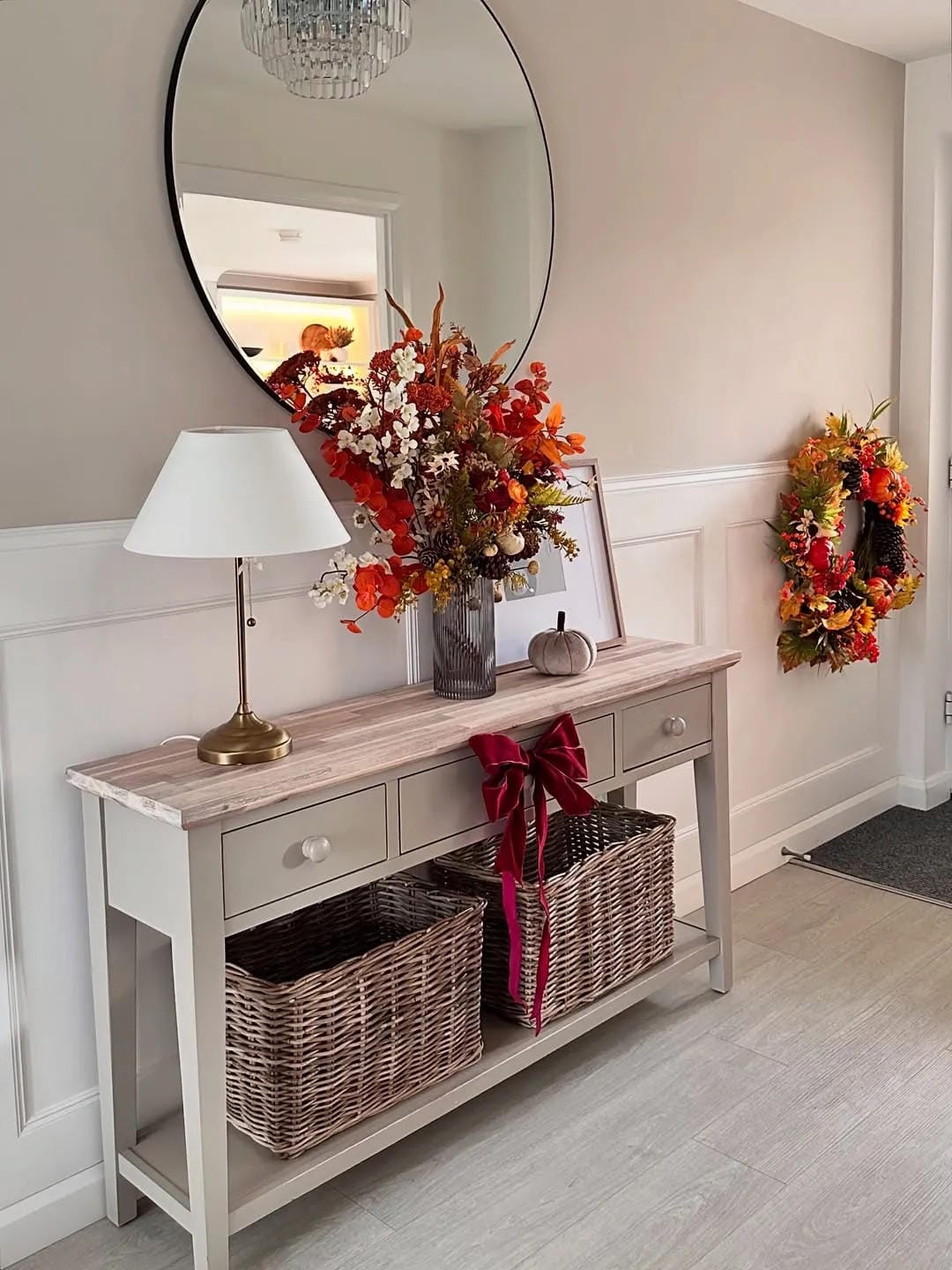 fall entryway table decor ideas with white console, fall flowers, matching fall wreath, lamp, basket, and round mirror