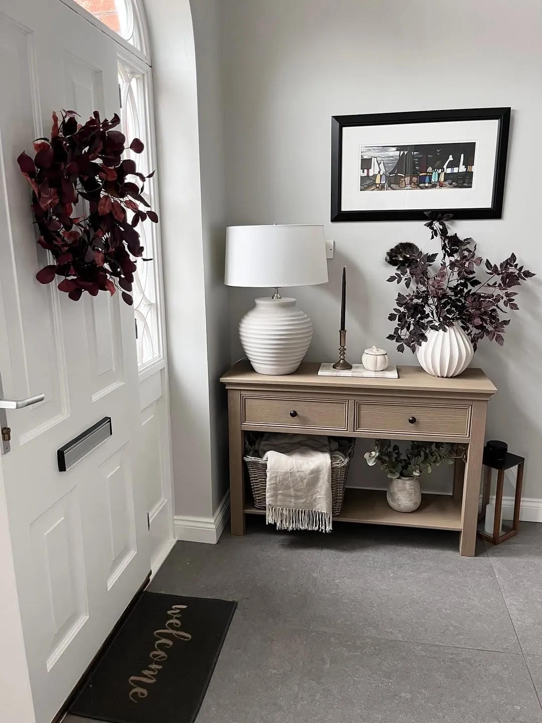 fall entryway table decor ideas with framed art, mirror, lamp, flowers, basket, and burgundy fall wreath on wall
