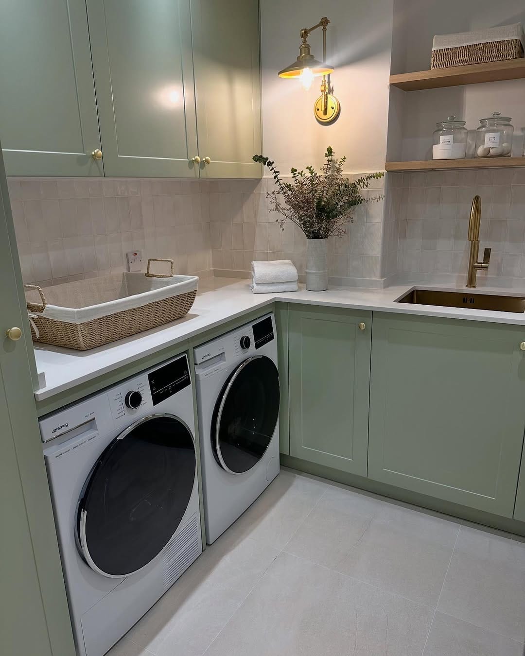 small laundry room ideas with green cabinets, brass wall light, open shelves, glass jars, basket, sink, and washer dryer