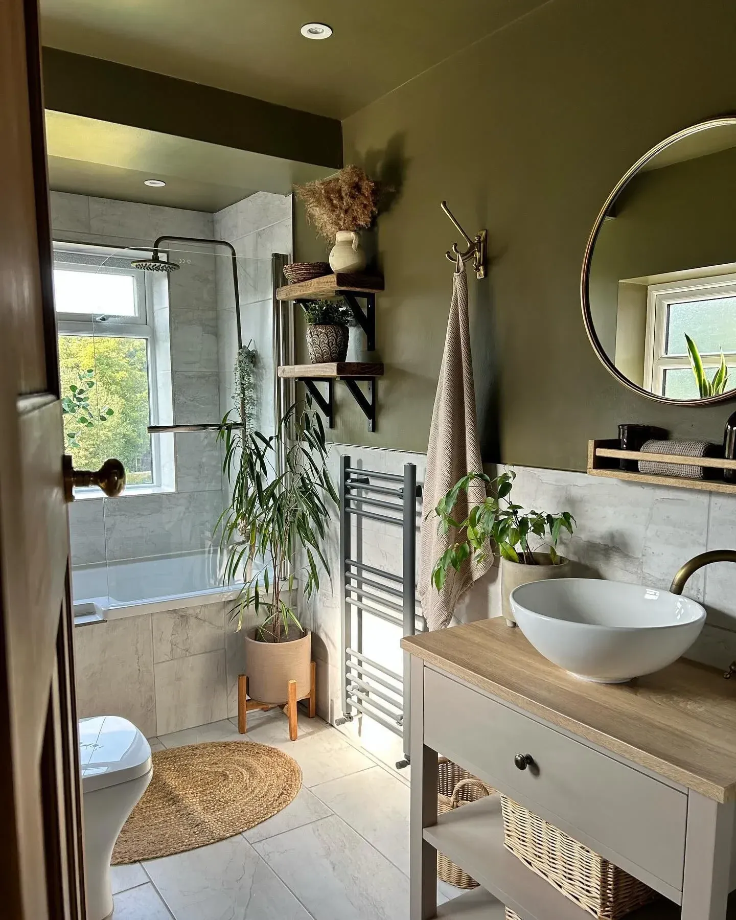 green bathroom ideas with olive walls, hanging plants, round mirror, wood vanity, and woven basket