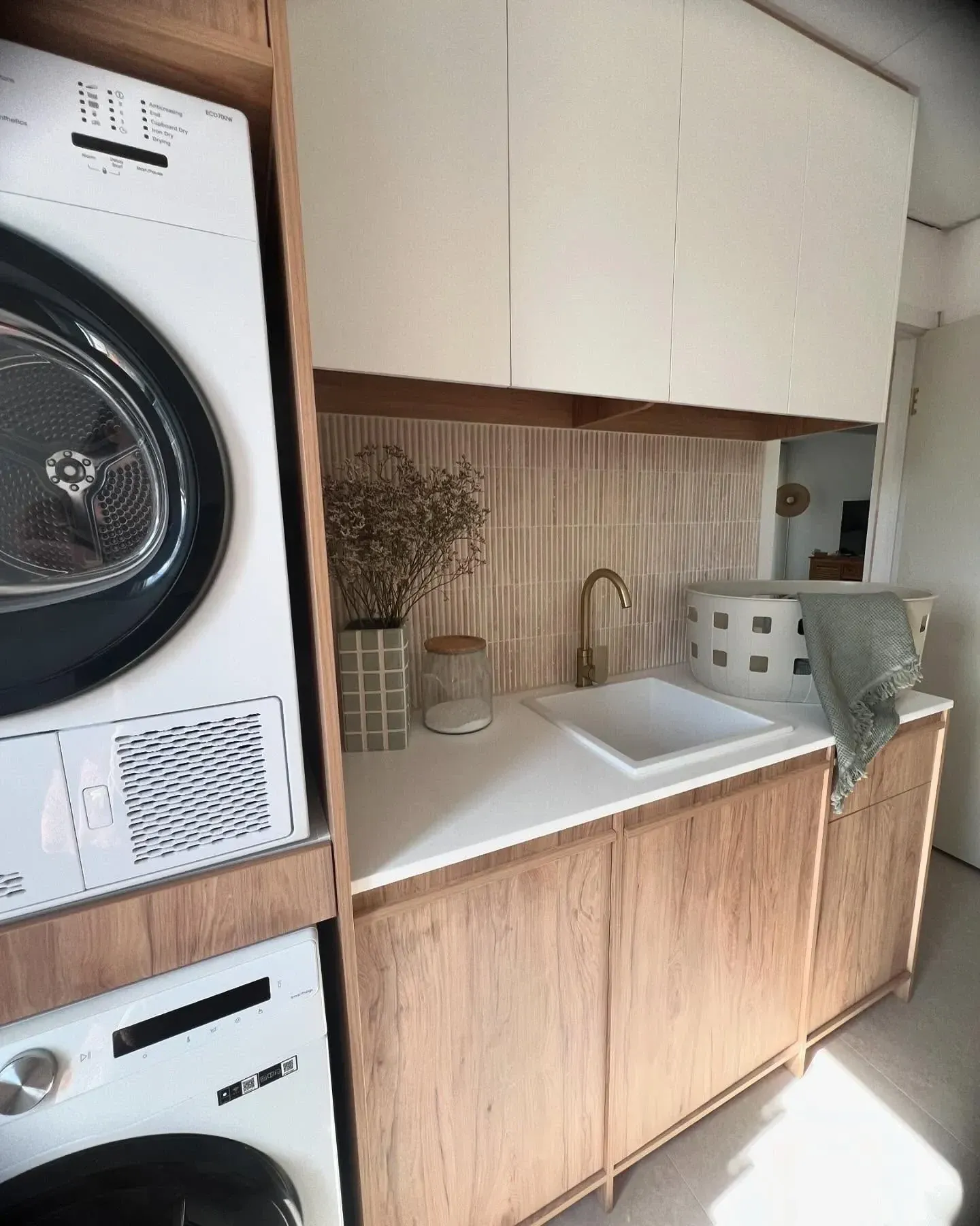 small laundry room ideas with stacked washer dryer, wood cabinets, white upper cabinets, sink, counter, and laundry basket