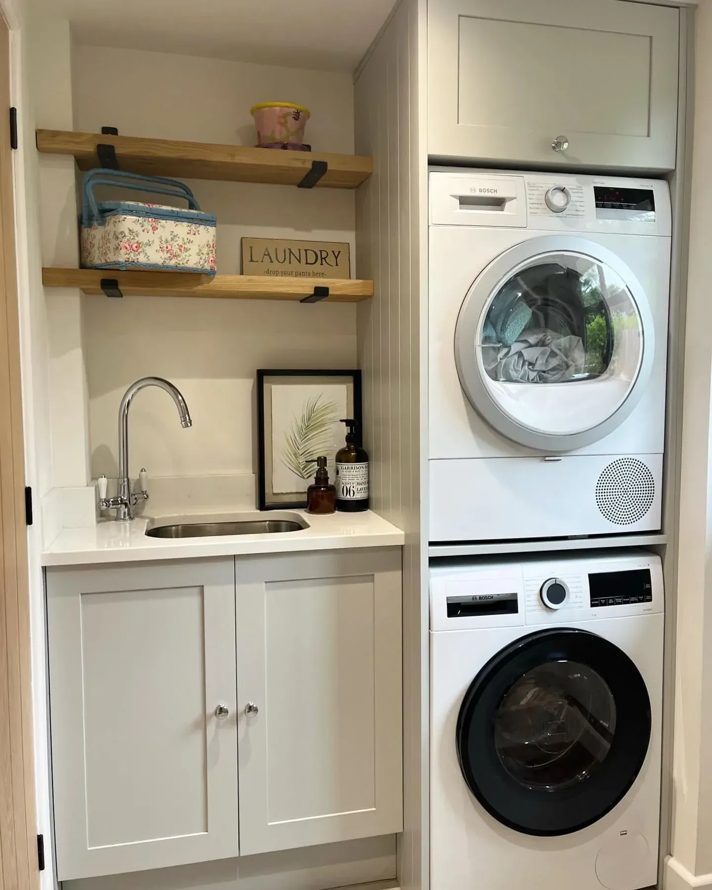 small laundry room ideas with stacked washer dryer, sink, open wood shelves, floral basket, and laundry sign