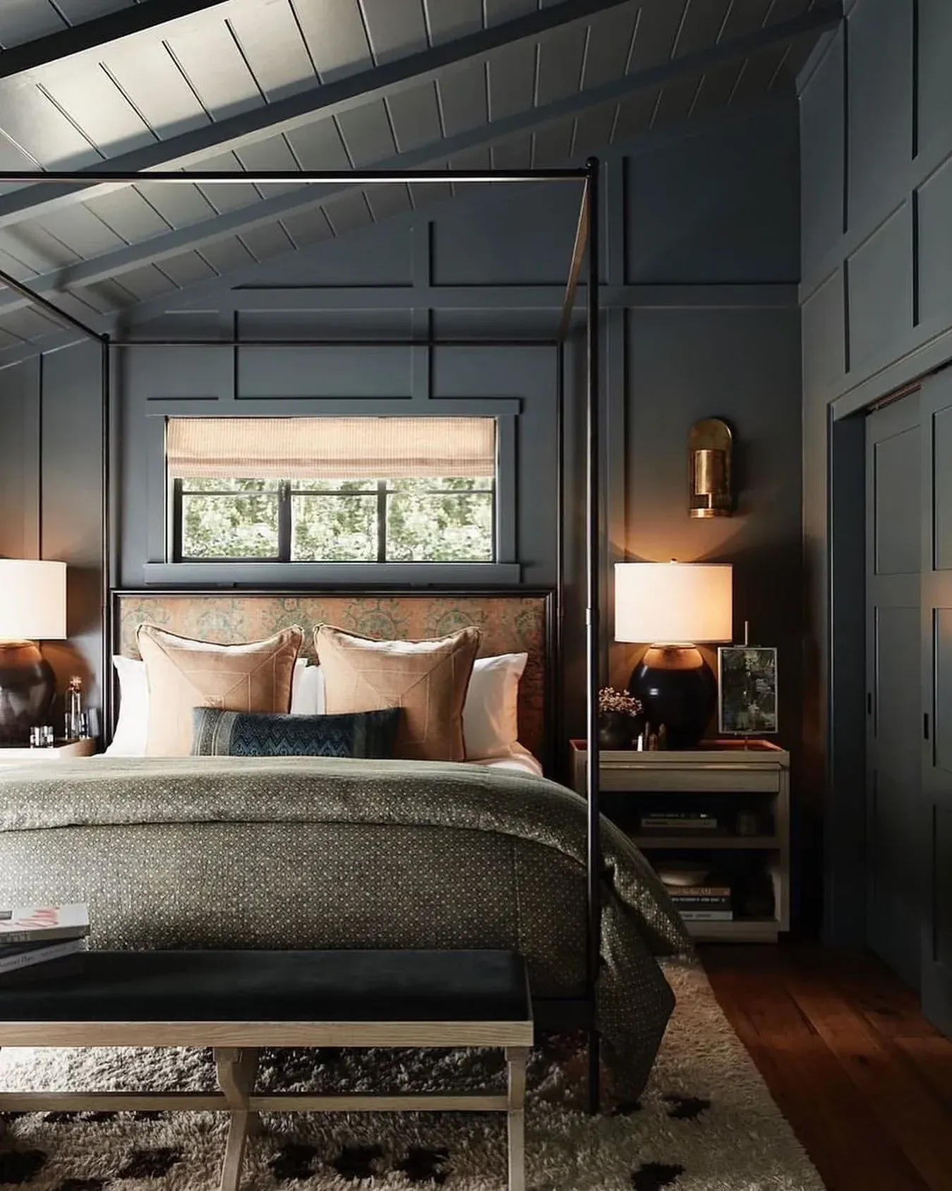 moody bedroom ideas with dark paneled walls, dark ceiling, warm lamps, bench, and layered bedding