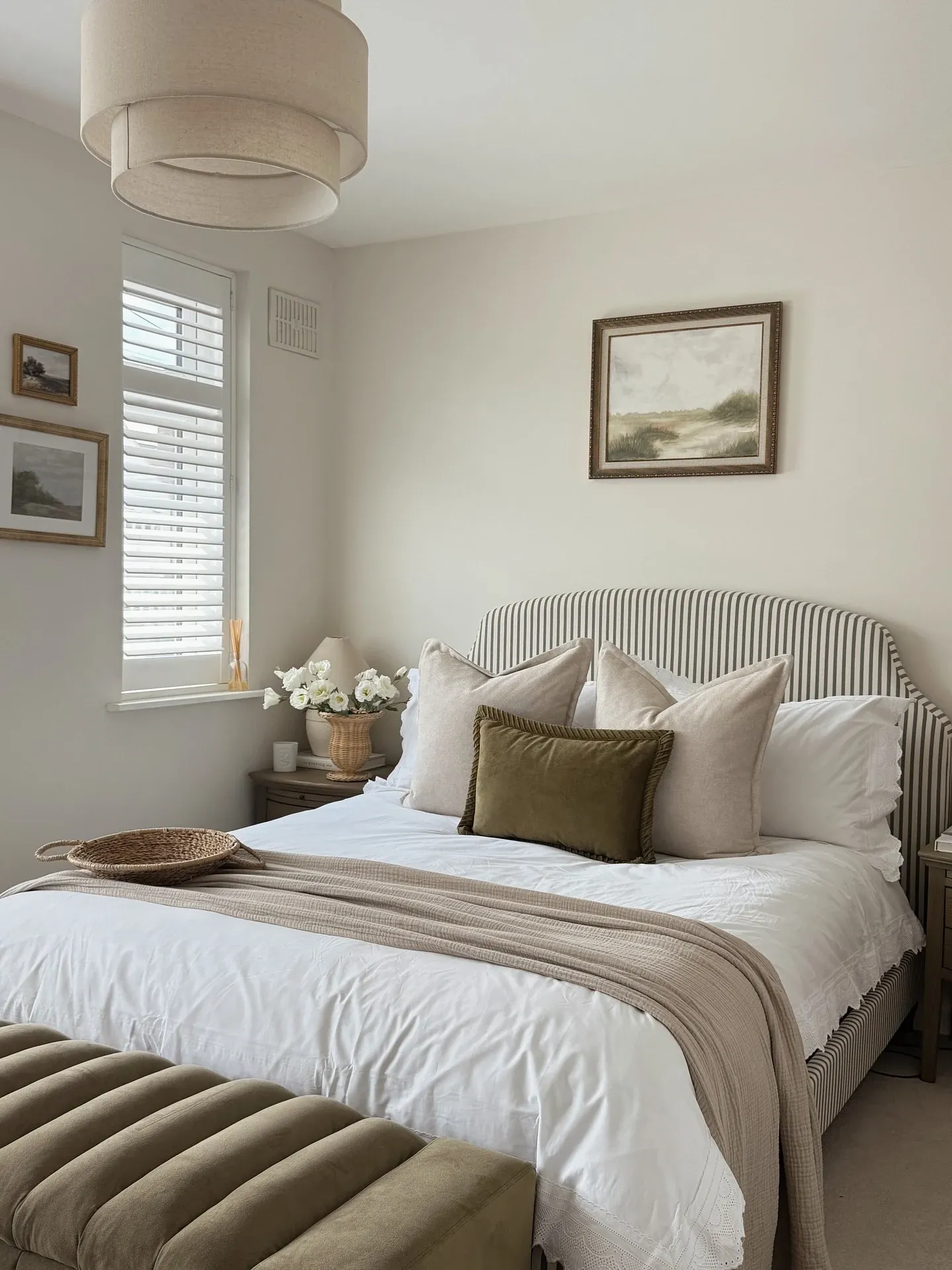fall bedroom ideas with olive pillow, white bedding, striped blanket, wood nightstand, woven bench, and soft natural light