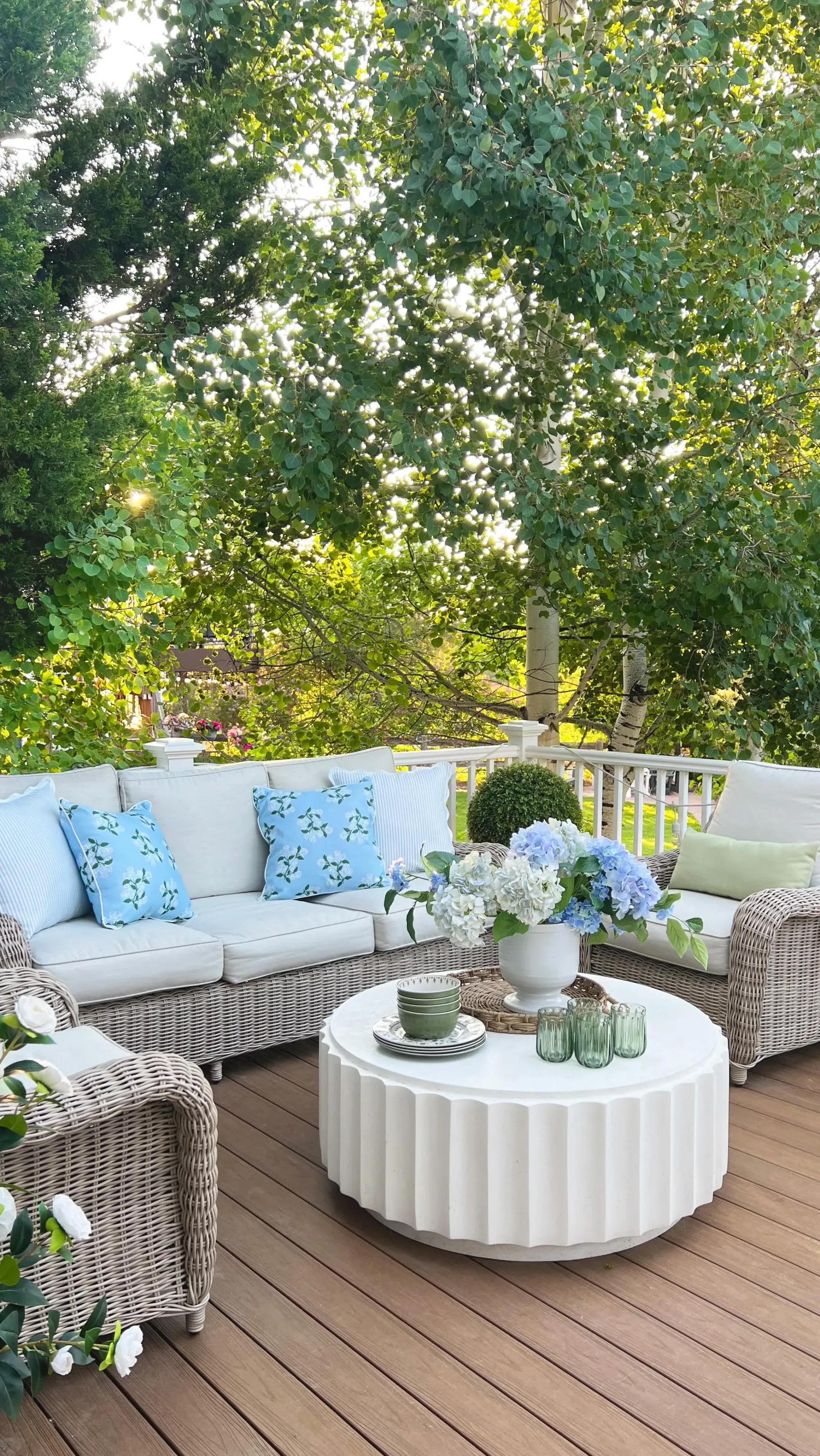 summer back porch decor ideas with wicker seating, blue pillows, round coffee table, flowers, and leafy backyard view