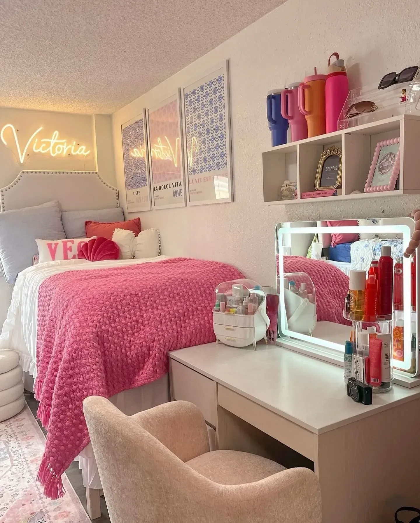pink and blue dorm room ideas with pink desk chair, vanity mirror, neon sign, pink bedding, blue wall art, and shelves