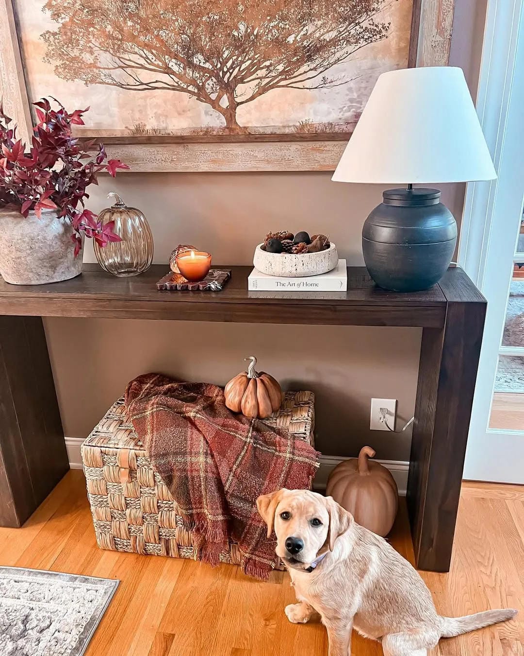 fall entryway table decor ideas with plaid blanket basket, pumpkins, lamp, candle, branches, books, and dog by console