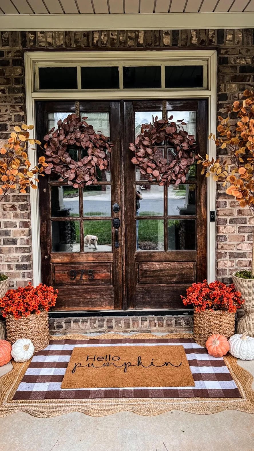 fall front door decor ideas with double wood doors, matching wreaths, pumpkins, fall flowers, and checked rug